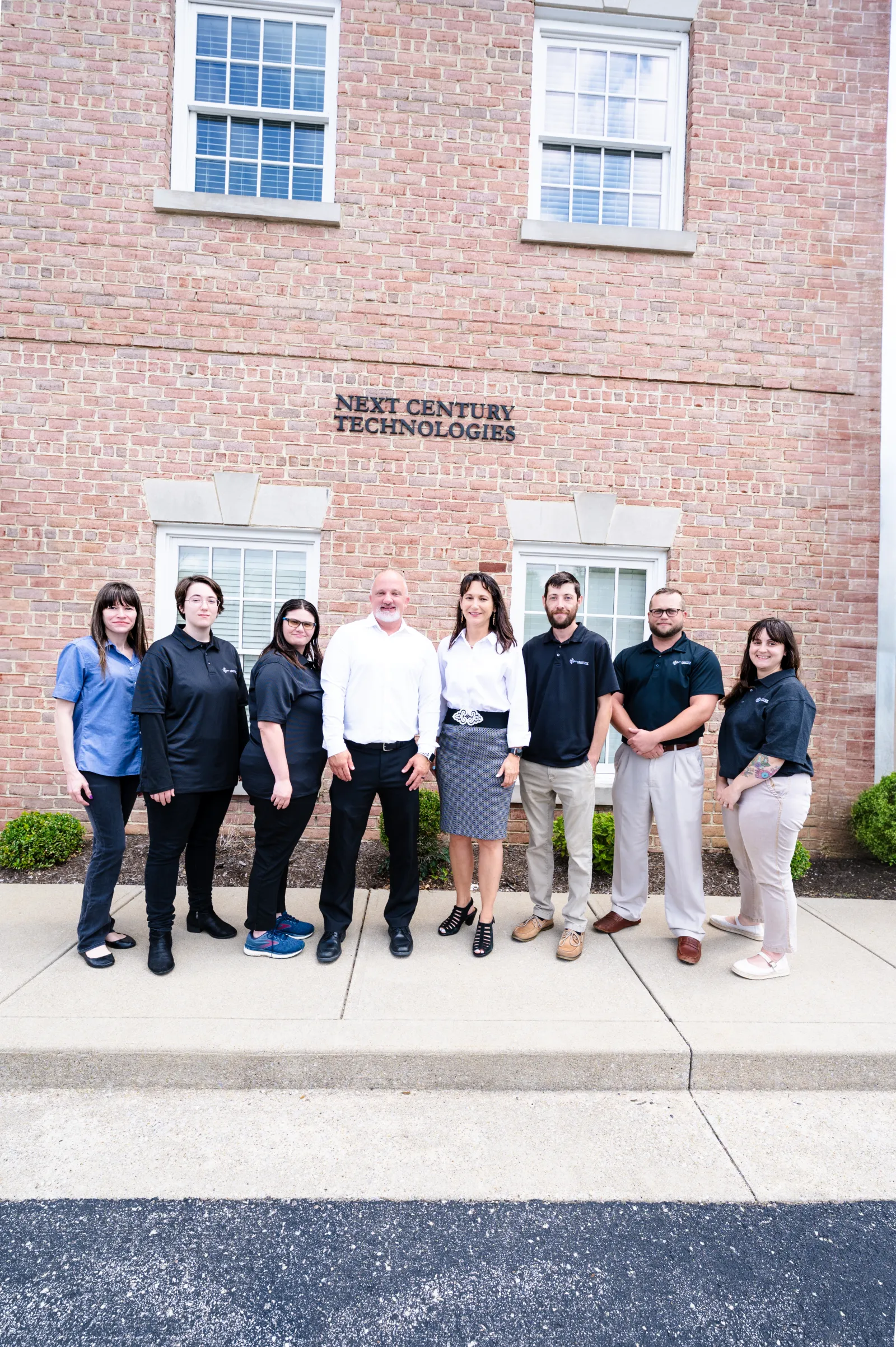 Eight professionals standing in front of Next Century Technologies office building with brick facade and windows.