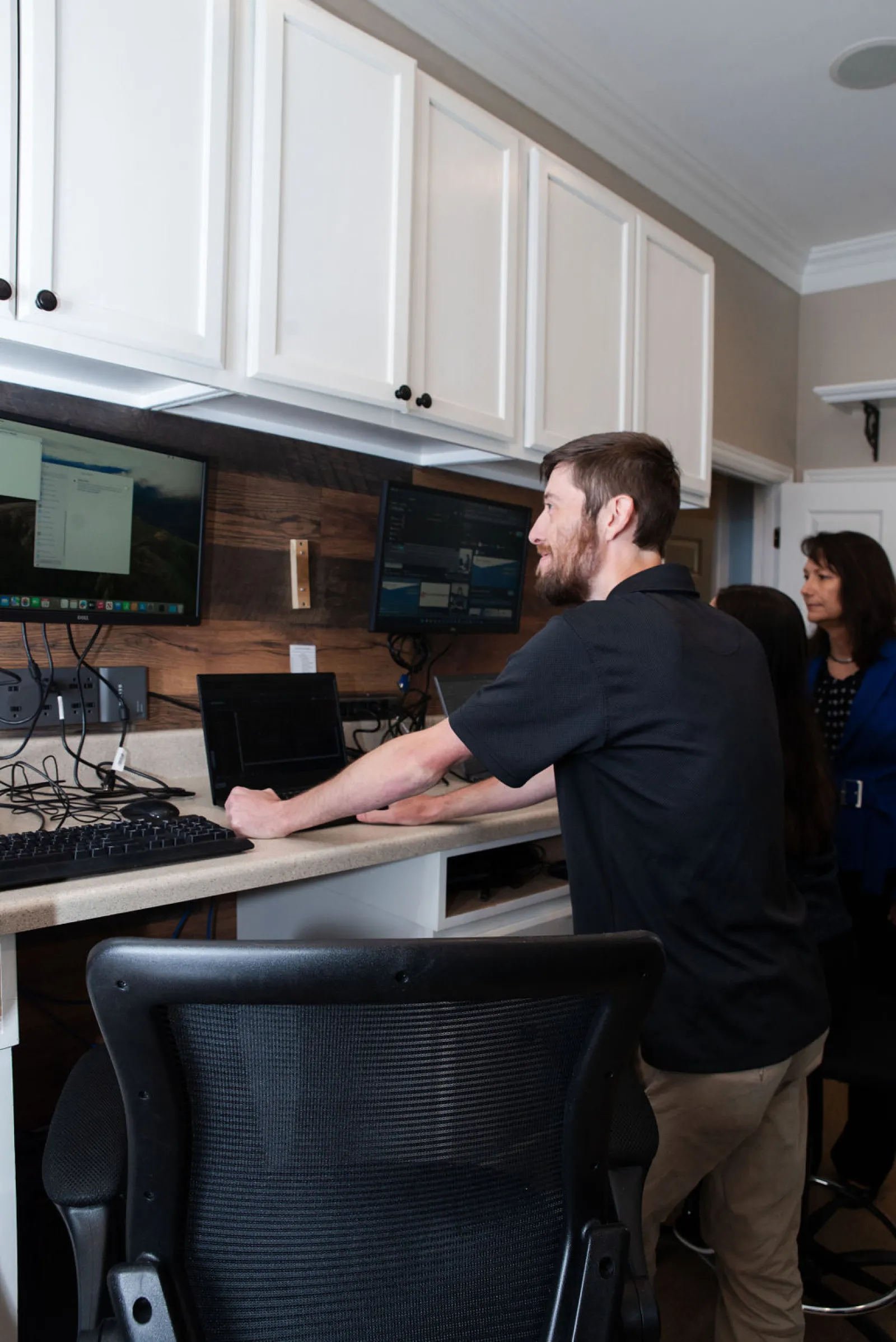 Man interacting with dual monitors and a keyboard in a modern workspace while two women observe.