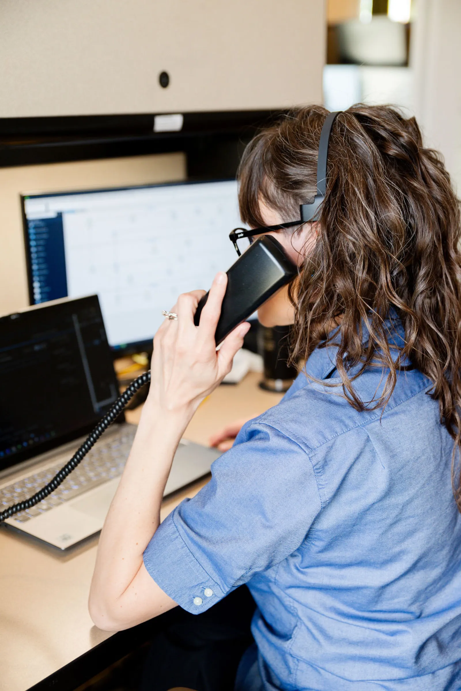 Woman in blue shirt speaking on desktop phone with headset, working on laptop and computer screens in office.