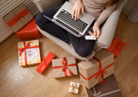 Image of pleased nice woman holding credit card with copy space using laptop while sitting on armchair surrounded by gift boxes at living room